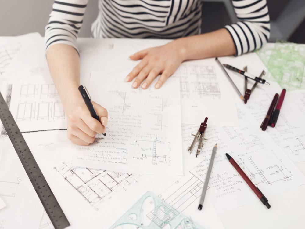 top-view-young-good-looking-freelance-engineer-wearing-formal-striped-clothes-working-comfortable-bif-table-making-notes-near-blueprints-fix-them-later-1-image